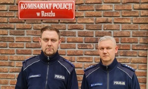 dzielnicowi od lewej asp. Paweł Ziółkowski oraz asp. szt. Paweł Obolewicz w tle czerwona tablica z białym napisem Komisariat Policji w Reszlu