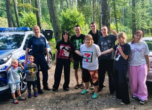 dzielnicowa podczas spotkania z dziećmi na półkolonii, grupa dzieci stojąca przy policjantce obok radiowóz w tle las