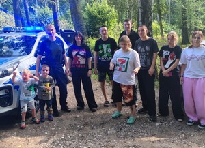 dzielnicowa podczas spotkania z dziećmi na półkolonii, grupa dzieci stojąca przy policjantce obok radiowóz w tle las