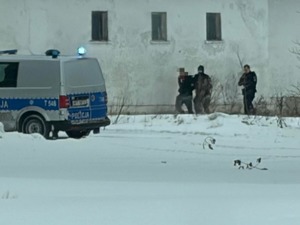 Policjanci prowadzą zatrzymanego mężczyznę obok radiowóz policyjny, w oddali budynek