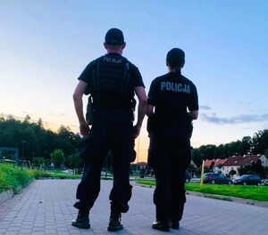policjant i policjantka stojący tyłem w oddali pojazdy i plac zabaw